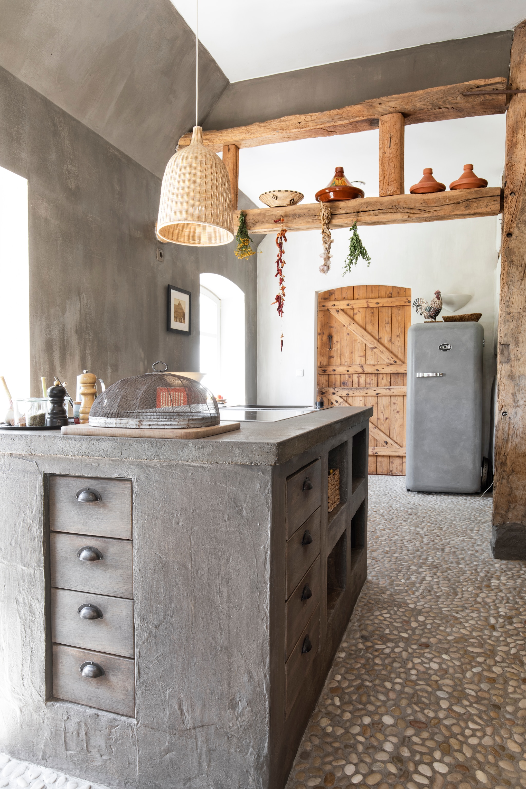 Hanglamp Leran van Ikea. Lades keukeneiland maatwerk. Koelkast in betonlook van Vintage Industries. Oude staldeur gezandstraald. Vloer Pebble Shop. vtwonen Landelijk 07-2026 | Styling Miriam Hannemann | Fotografie André Reuter