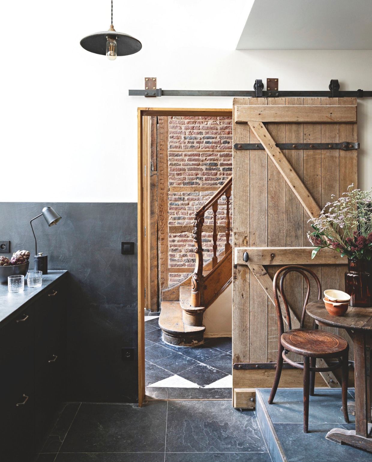 Keuken beneden. Vloer, bakstenen muur en trap in de hal zijn origineel. Keukenvloer is behouden van de laatste renovatie. Muurverf Velvet Satin in Not Totally White van Ressource. Lambrisering van en mdf werkblad bekleed met zwart, mat gepolijst beton door Marius Aurenti. Oude deur op rail van L’Herminette. Keukenkasten Ikea. Zwarte fronten Fenix van Bocklip. Handgrepen Vintage. Quincaillerie Leclercq. Hanglamp Studio Racines. Tafellamp Am.pm. Slakom La Redoute. Waterglazen Monoprix. Stoel en tafel Selency. Terracotta kommen Doux Août. Vaas Leboncoin. vtwonen Landelijk 04-2026 | styling Marie-Maud Levron | fotografie Yann Deret