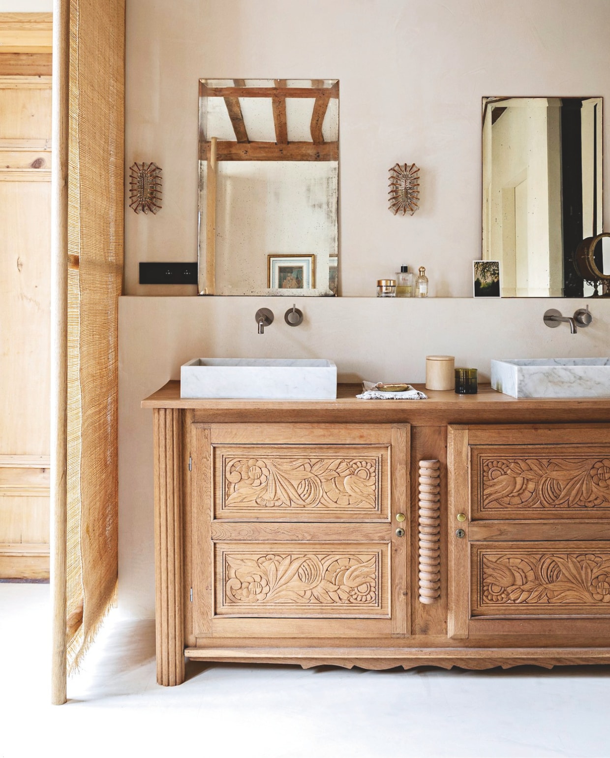 De badkamer en suite wordt van de slaapkamer gescheiden door eiken-houten rekken van Cdr Sophie bedekt met stroken raffia van Adèle Collections. Vloer, muren en plafond bedekt met gepolijst beton in kleur Parchemin door Marius Aurenti. Wastafelmeubel, spiegels en glazen wandlampen Selency. Waskommen Leboncoin. Mengkranen Gessi. vtwonen Landelijk 04-2026 | styling Marie-Maud Levron | fotografie Yann Deret