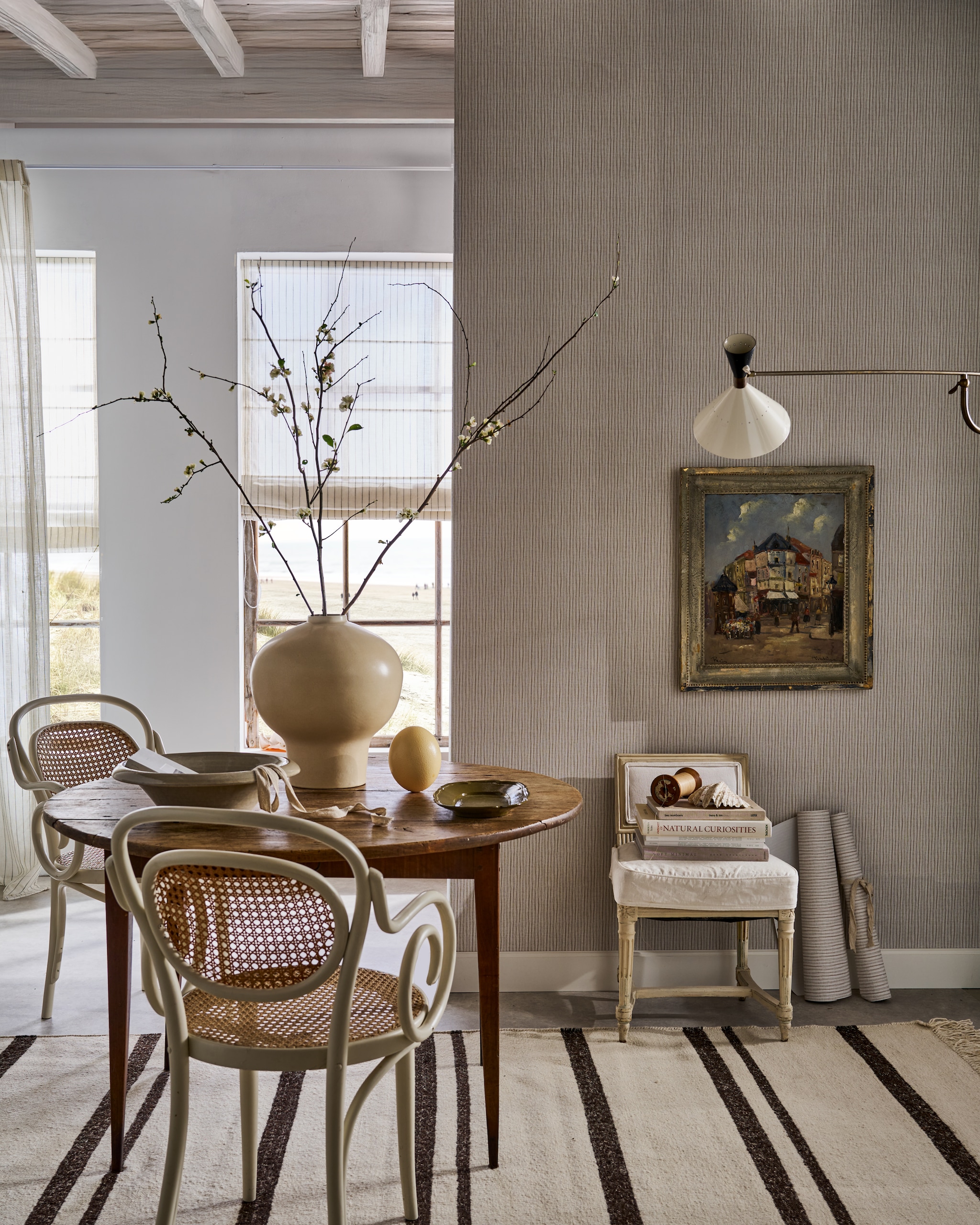 vtwonen behang Double Stripes, vliesbehangpapier, warm grey, 10 m x 53 cm (lxb), per rol € 35,99.M.m.v. prinsheerlijkantiek.nl (schilderij, tafel, gestoffeerde stoel), hkliving.com (vloerkleed), urbannatureculture.com (vaas), karwei.nl (vouwgordijn, muurverf en gordijn van vtwonen), pietjonker.nl (wandlamp). zarahome.com (schaal).Overig privébezit.