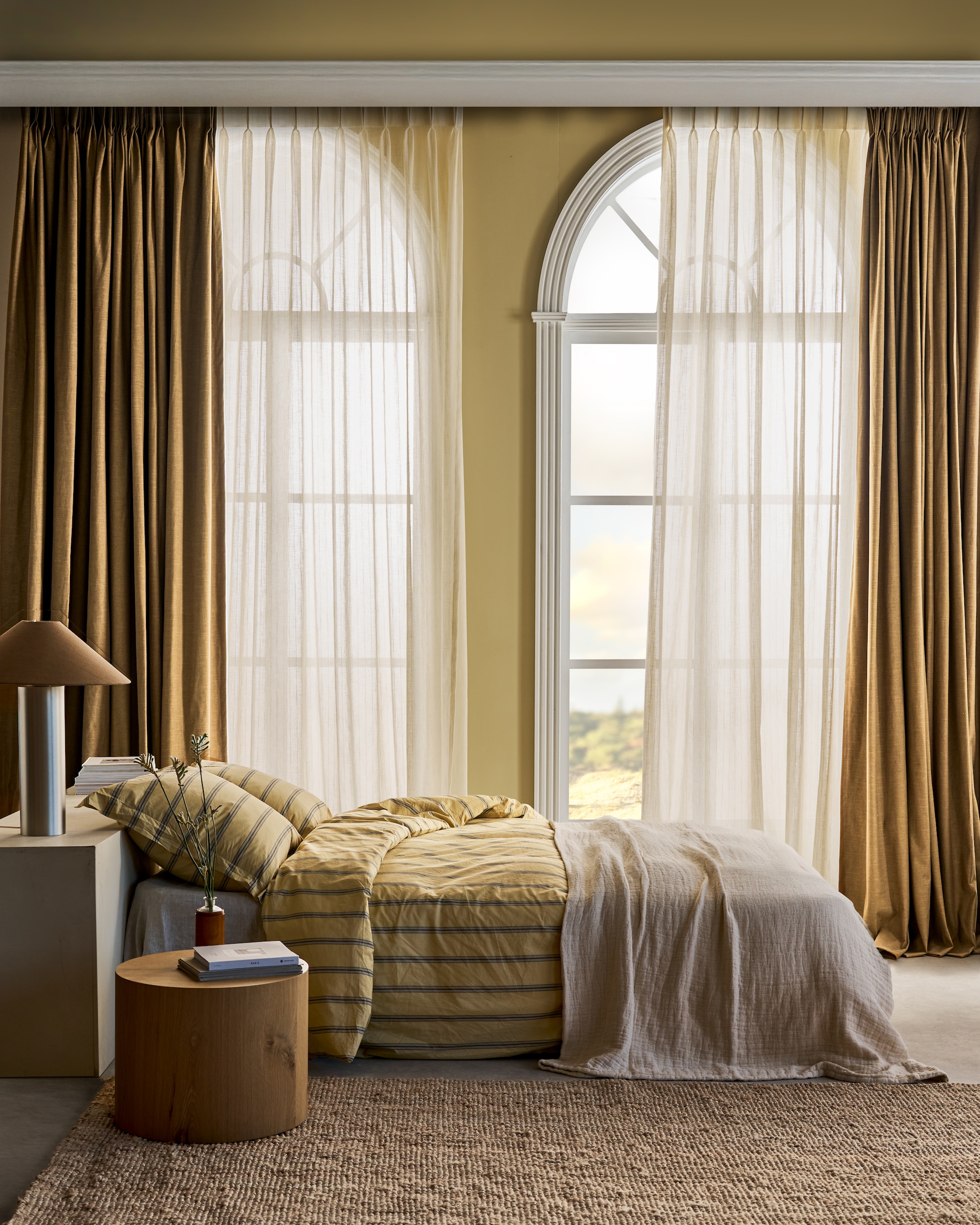 vtwonen transparant gordijn op maat Wide Stripe, kamerhoog, ecru, per strekkende meter vanaf € 29,99, vtwonen verduisterend gordijn op maat Linen Mix, mustard, per strekkende meter vanaf € 38,99. M.m.v. zuiver.com (bijzettafel), zarahome.com (dekbedovertrek, sprei, vloerkleed), karwei.nl (verf kleur wax yellow). vtwonen 07-2026 | styling Cleo Scheulderman | fotografie Jeroen van der Spek