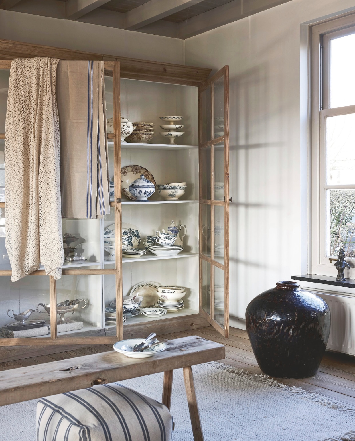 Het Kabinet Wonen kast tweedelig xl, grenen, naturel, 220x50x231 cm (bxdxh) € 2795,-, bank, hout, naturel, 182x37x44 cm (bxdxh) € 195,-, vloerkleed Woolie, wol, roomwit, 290x200 cm € 499,-, pot, aardewerk, bruin € 395,- (hetkabinet.nl). vtwonen poef, katoen en linnen, beige € 69,95 (shop.vtwonen.nl). Tableware Amsterdam borden klein, keramiek, wit en blauw, per stuk € 25,-, borden diep, keramiek, wit en blauw, per stuk € 35,-, borden groot, keramiek, wit en blauw, per stuk € 35,-, taartplateau, keramiek, wit en blauw € 40,-, schalen rond, keramiek, wit en blauw, per stuk € 40,-, platte schalen ovaal, keramiek, wit en blauw, per stuk € 175,-, juskommen, keramiek, wit en blauw, per stuk € 25,-, soepterrines, keramiek, wit en blauw € 95,- en € 125,-, kop en schotel, keramiek, wit en blauw € 10,-, theepot, keramiek, wit en blauw € 45,-, bestek, zilver, per stuk € 5,-, tafelkleed, linnen, beige en blauw € 75,-, beeld, kunstenaar A. Nelson, ca. 1900, brons, donkergrijs € 650,- (kramer.amsterdam). Schaal op voet met oor, metaal, verzilverd € 80,-, schalen, metaal, verzilverd € 75,-, juskom, metaal, verzilverd € 80,-, botervloot, metaal, verzilverd € 45,-, schaal op voet, metaal, verzilverd € 75,-, eendenhoofd, metaal,  verzilverd € 65,- (tableware.amsterdam).  vtwonen landelijk 6-2026 | Styling Marianne Luning | Fotografie Alexander van Berge