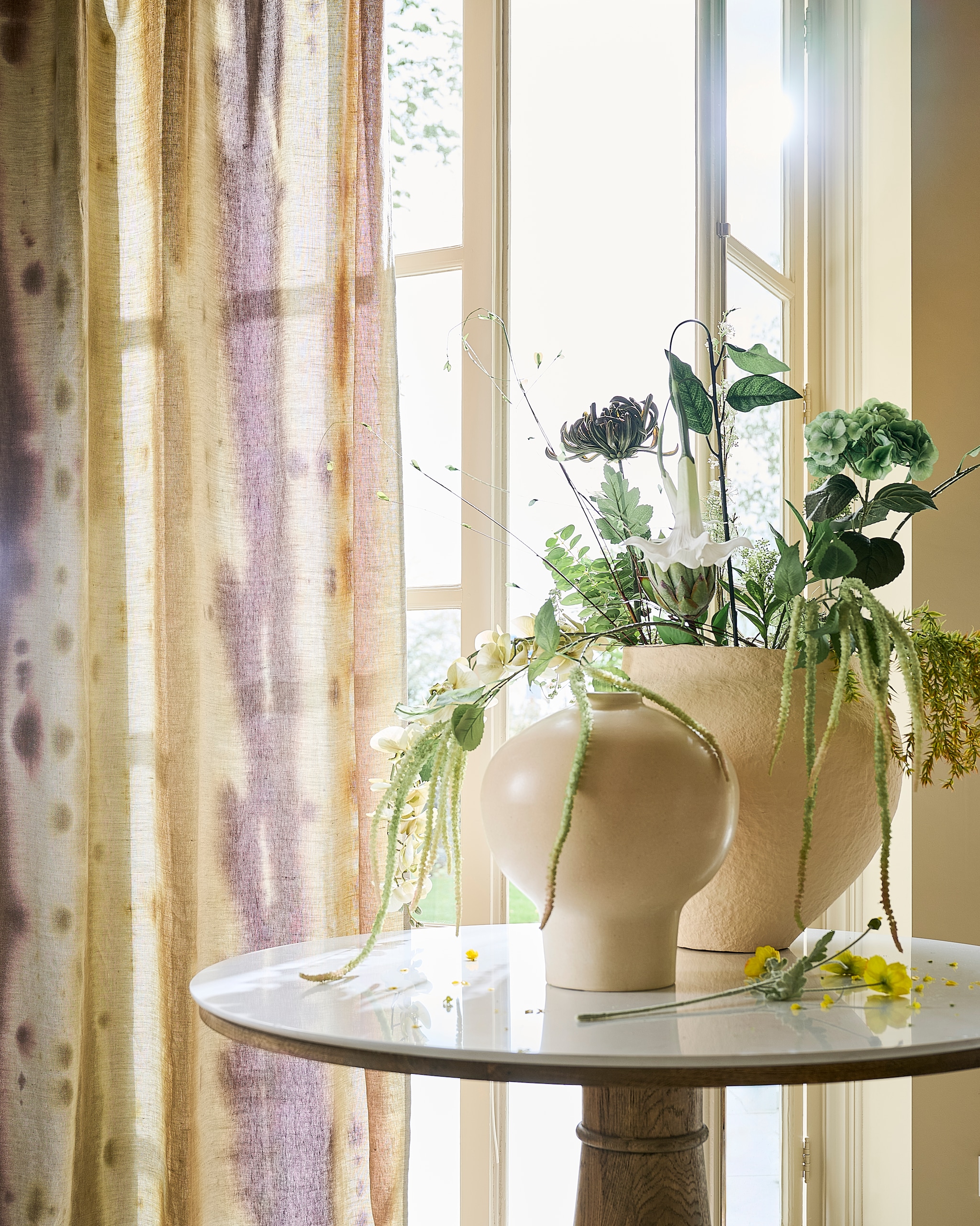 Designers Guild gordijnstof Brera, linnen, naturel, geel en bruin, per meter € 169,- (wva.nl). Rivièra Maison tafel Amalfi, steen en hout, wit en bruin, 130x76 cm (Øxh) € 1799,- (rivieramaison.com). Urban Nature Culture pot Sandia, katoen maché, off white € 249,-, pot Ama, keramiek, offwhite € 169,99 (shop.vtwonen.nl). Atelier Verden kunstbloemen, kunststof, groen, vanaf € 12,50 (atelierverden.com).