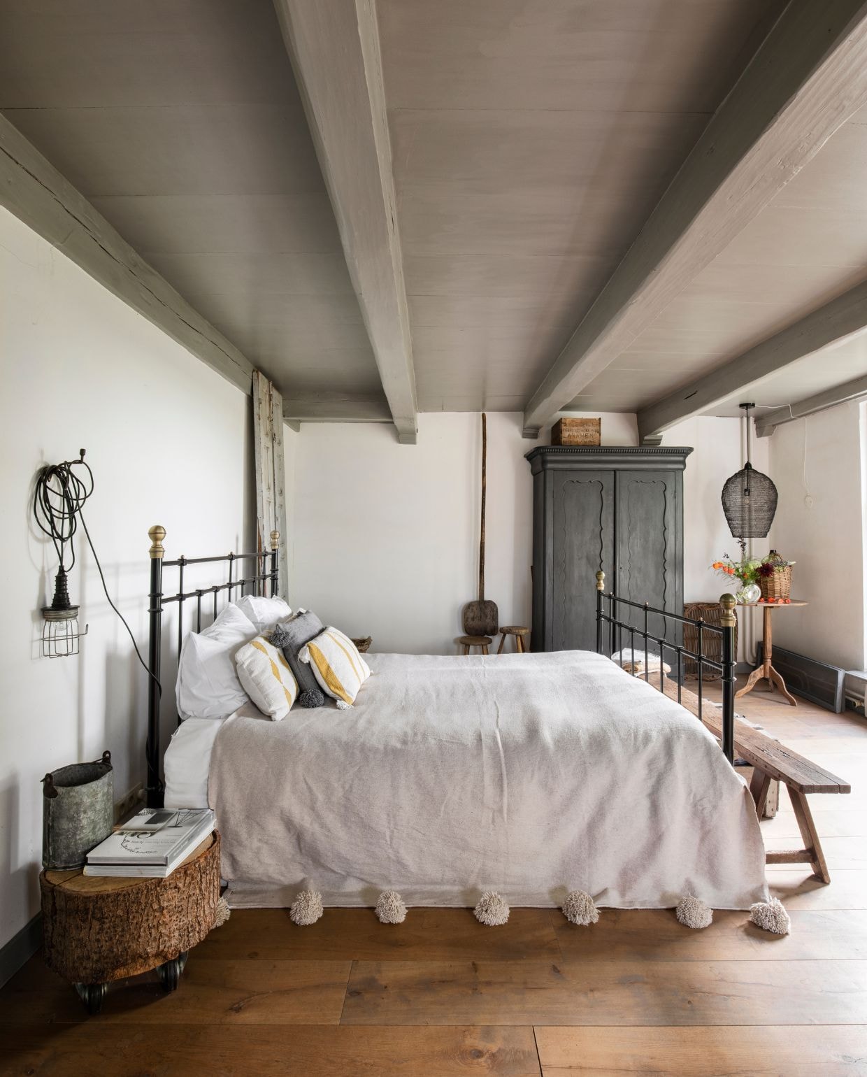 Bed Marktplaats. Sprei The Souks. Nachtkastje gemaakt van een stuk boomstam. Looplamp uit werkplaats. Kast gevonden in de schuur en grijs geverfd. Bank brocante. Kleur plafond betongrijs Ral 7023. vtwonen Landelijk 05-2026 | styling Miriam Hannemann | fotografie André Reuter