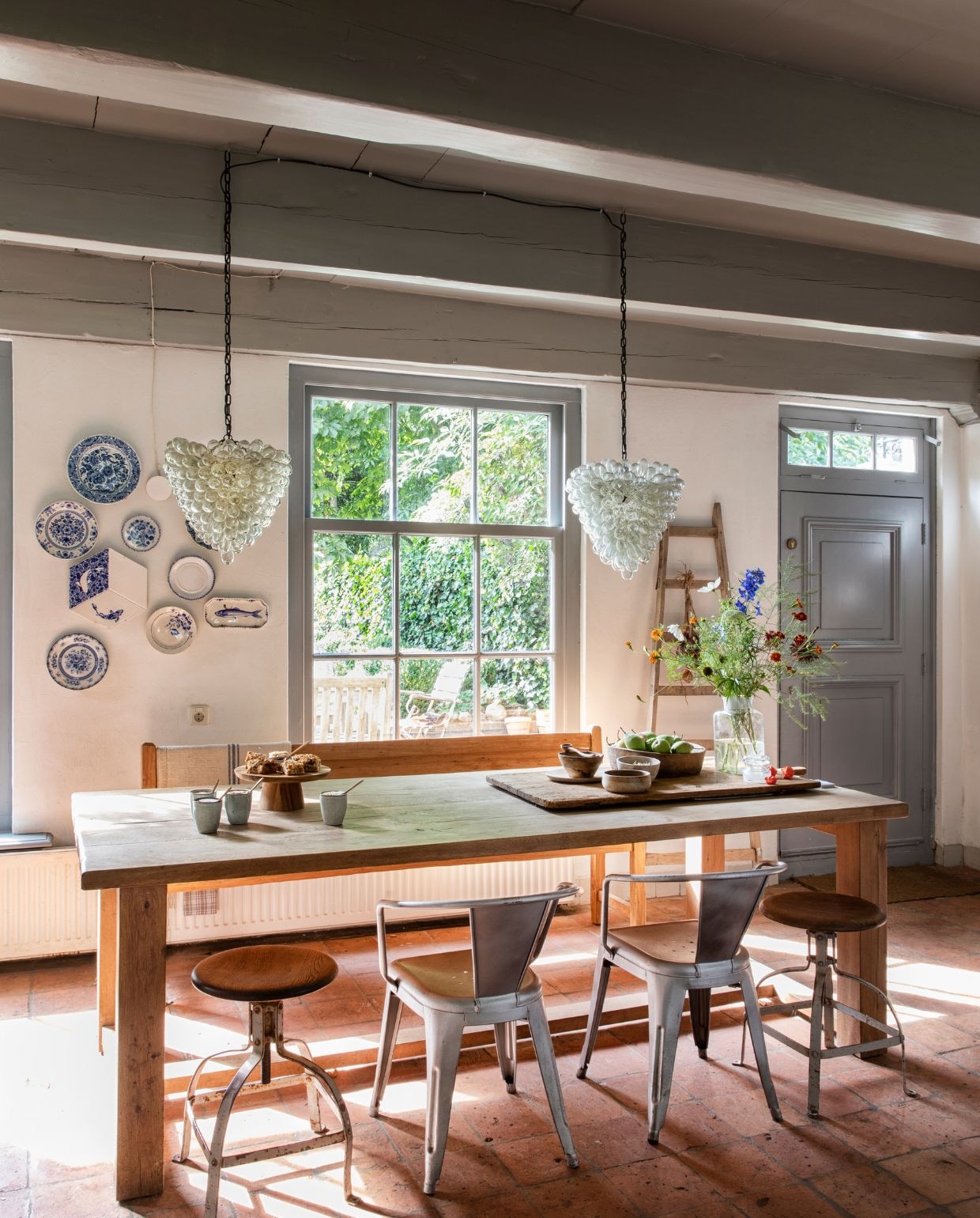 Eettafel Pilat&pilat. Metalen stoelen HKliving. Plavuizen uit de tijd dat hier de bakkerij zat. Krukken brocante. Verzameling Delfts blauw antiek en van ontwerpers Tichelaar Harlingen en Heinen Delft. Hanglampen via Marktplaats. vtwonen Landelijk 05-2026 | styling Miriam Hannemann | fotografie André Reuter