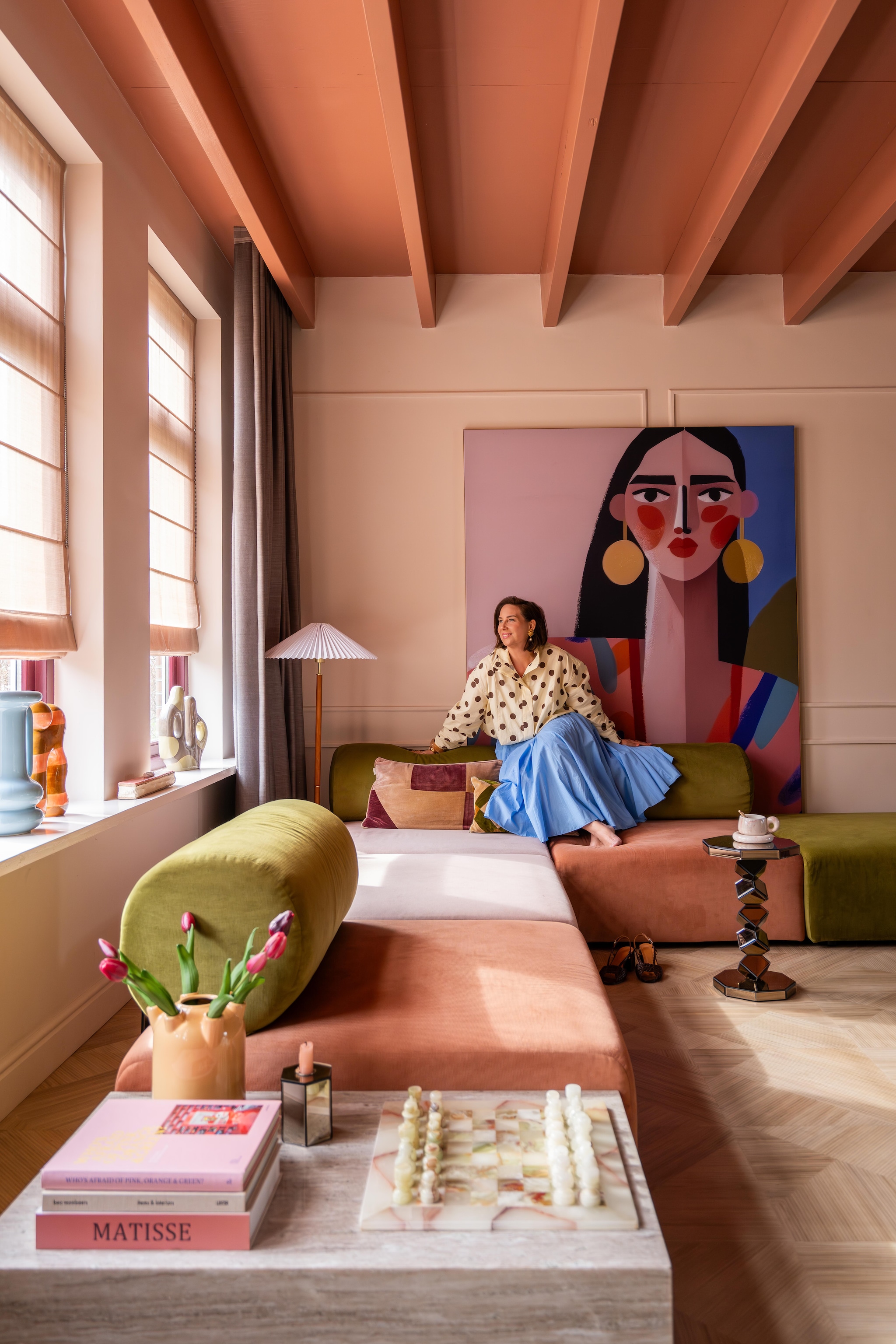 Bank Zitmaxx. Schilderij Carla Van Iersel via Werk aan de Muur. Staande lamp H&m Home. Blinds Kendix. Gordijnen De Ploeg. Vloer Moduleo. Hoektafels, blauwe vaas en spiegeltafel By-Boo. Schaakbord The Vintage Atelier. Kandelaars Kwantum. Kussens Sézane. Verfkleuren plafond Light Cherry Parade en wand Peach Pudding van Histor.