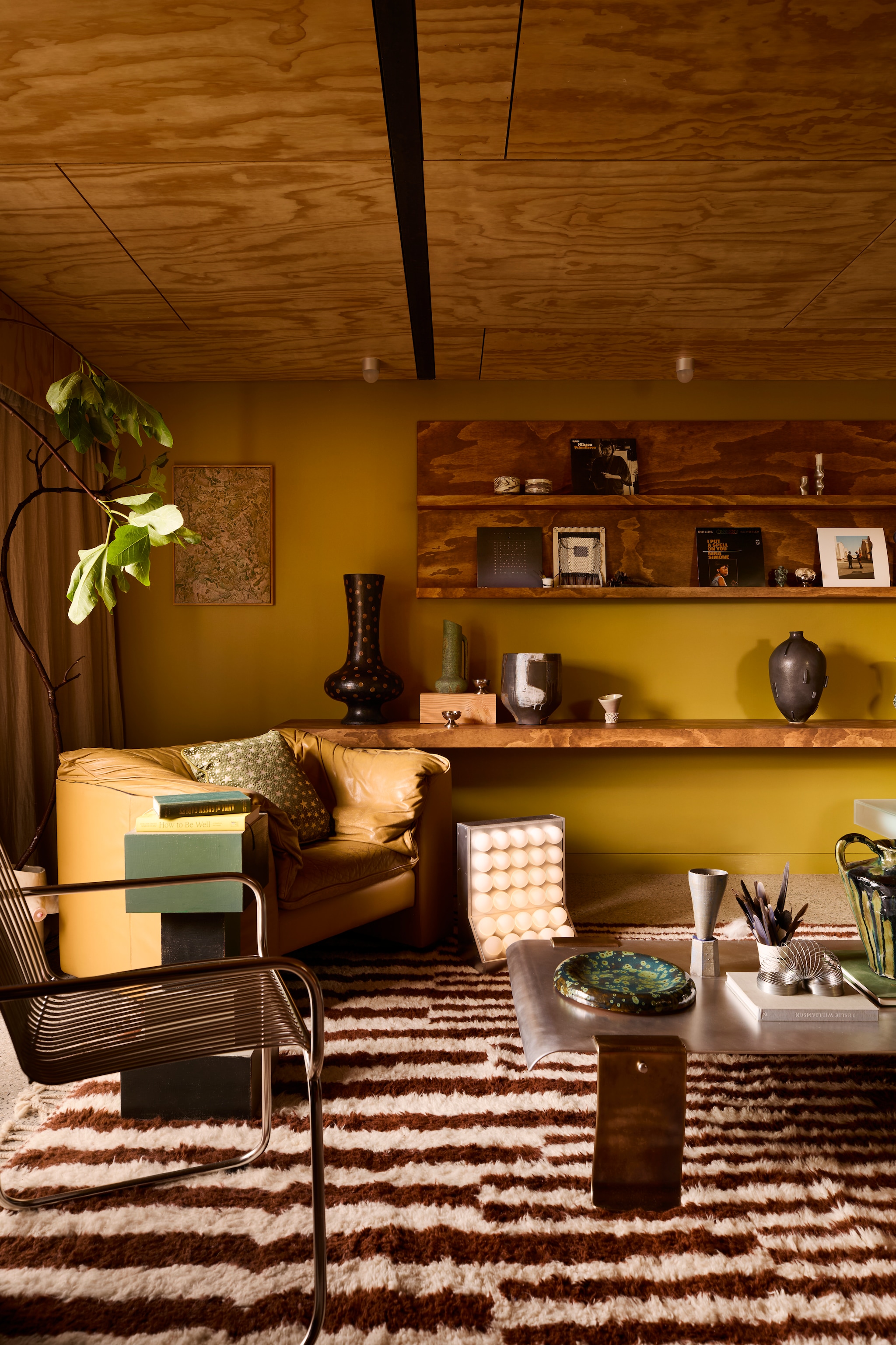 Muurkleur Plantation Island van Dulux. Radiator Ply van Maxiply. Plafondlampen Objectsforthought. Kunstwerk links Ro Noonan. Fauteuil vintage uit de jaren 80. Kussen Celeste van Tali Roth. Salontafel Jordan Fleming. Op salontafel: bord Mossy Platter van Rina Bernabei, object Trophy #2 van Alterfacts en sculptuur Drip Urn van Kelly Brown. Vloerkleed Moroccan Shag van Afrikesh op Etsy. Bovenste wandplank: luciferkommetjes Fdo Studio, kandelaar Locki Humphrey. Onderste plank: donkergrijze vaas Astrid Salomon/Bastard Ceramics, hoge vaas Martyn Thompson, groene vaas Tantri Mustika Ceramics, pot Steph Wallace, object Trophy #4 van Alterfacts.