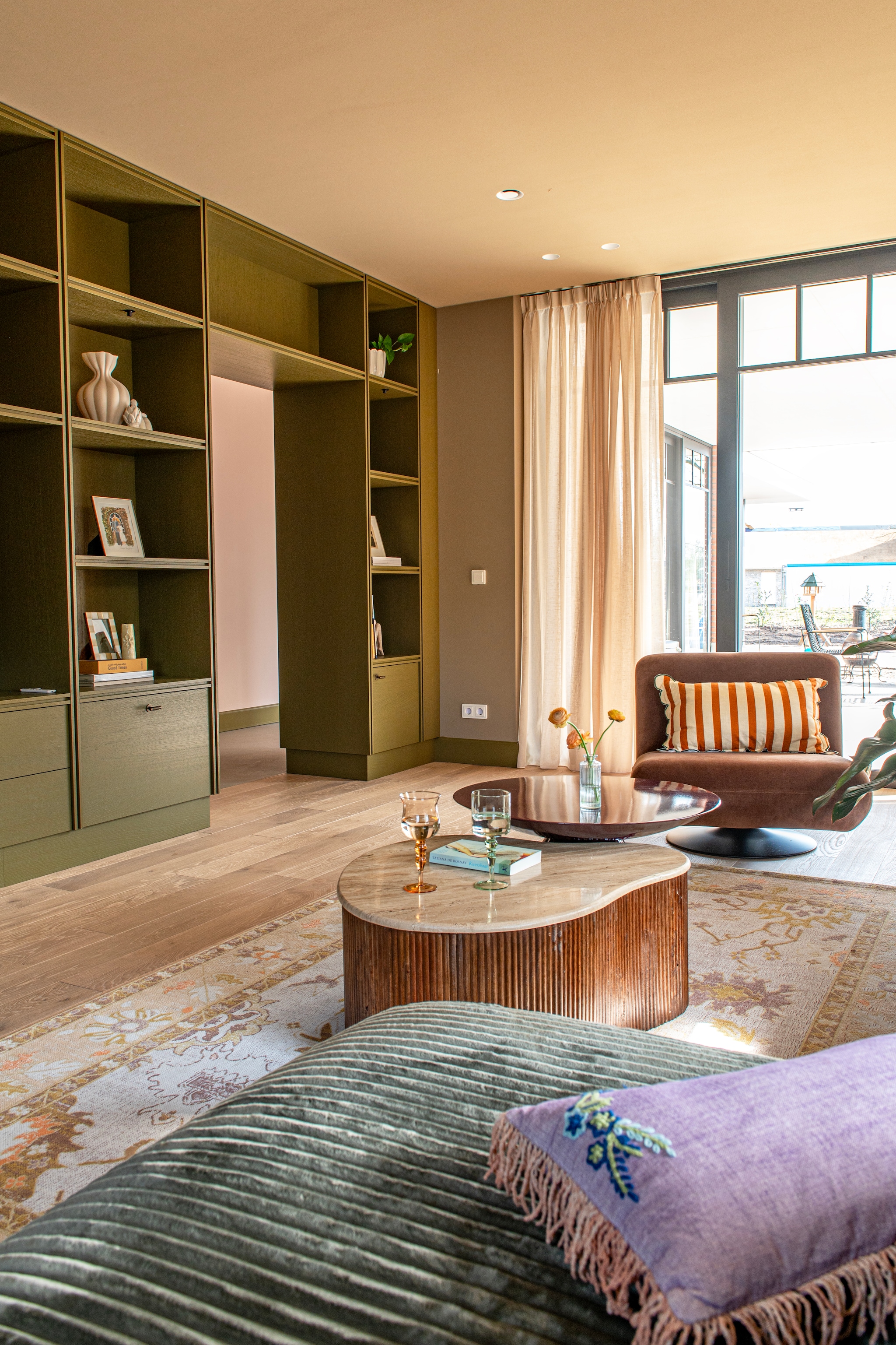 Maatwerk kasten geschilderd in Hunter van Lab Paint. Vouwgordijnen Gotain. Kleur op de muur zelf gemengd. Bank Micadoni. Stoel Zara Home. Salontafel met hout Giga Meubel. Ronde bijzettafel, rond roze kussen en vloerkleed Sissy-boy. Langwerpig kussen Zara Home.  Overige kussens Rice. Lamp HKliving. Wijnglazen Westwing. vtwonen Special 03-2026 | fotografie Caroline Coehorst | productie De Binnenkijkers by Caroline
