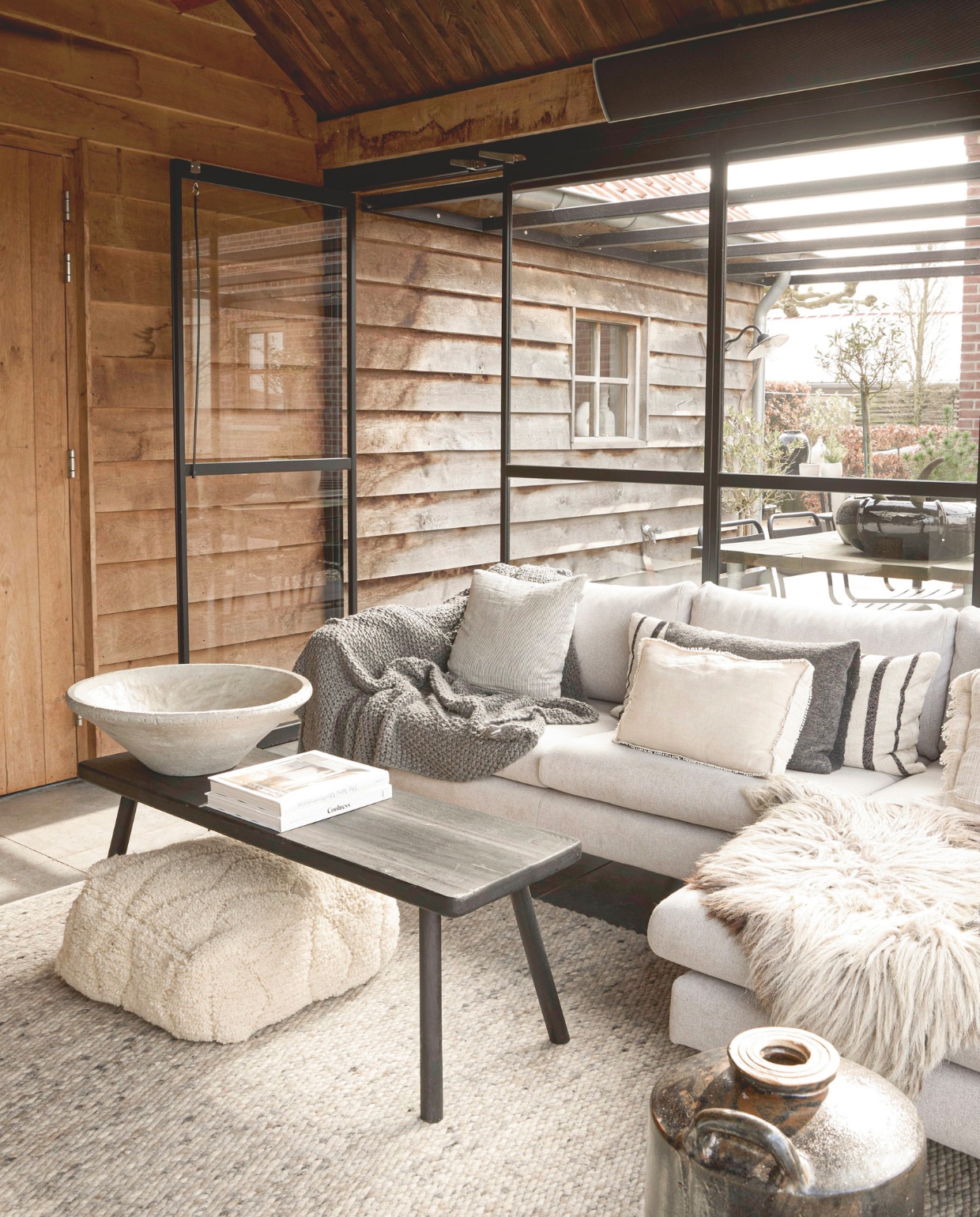 Stalen pui  Metalura. Bank Ikea. Keramische tegelvloer. Vloerkleed De Agave Natuurlijk Wonen. Sprei H&m. Texelse Schapenvacht. Salontafel, boeken en vierkante kussen,  De Tuynkamer. Kussens Katoo Fashion&living. Poef Studio Pure. Schaal en schaal buiten Snufenshoe. Kruik ’t Potterieke. Tafel buiten zelf gemaakt. Stoelen buiten Intratuin. vtwonen Landelijk 05-2026 | styling Kristel van Leeuwen | fotografie Peggy Janssen