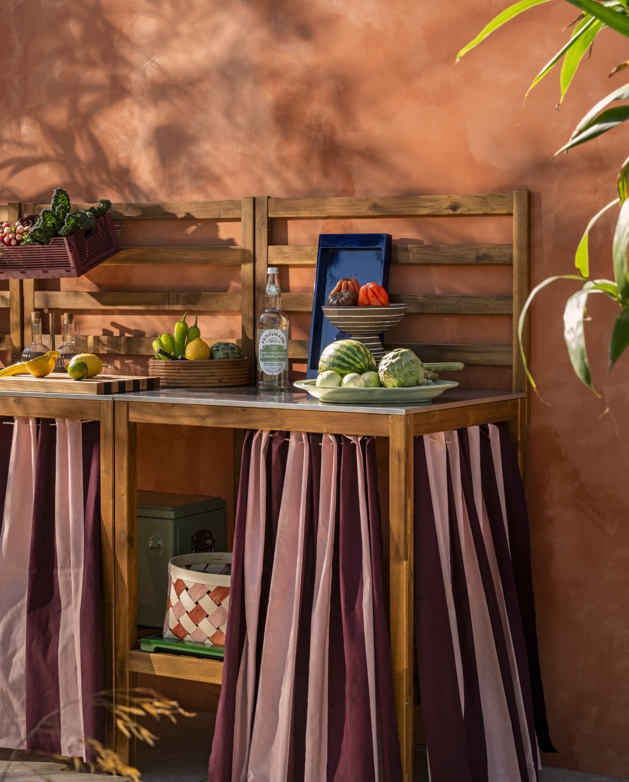 Ikea buitenkeuken Nämmarö, roestvrij staal en acaciahout, 240x62x140 cm (lxbxh) € 417,-, mand Ommjänge, gevlochten vezel, oranje en roze, set van 2 € 19,99 (ikea.nl). Arket verhoogde schaal, keramiek, groen, 22 cm (Ø) € 49,-, dienblad, gelakt hout, blauw€ 45,-, dienblad, gelakt hout, groen € 55,-, citruspers, kunststof en metaal, geel € 15,- (arket.com). Ferm living snijplank Stripe bold, walnoot- en eikenhout, naturel en bruin € 139,-, dienblad Isola, hout, naturel, set van 2 € 109,-, schaal op voet Brura, keramiek, bruin en wit € 59,- (fermliving.com). H&m Home tafelkleed, gebruikt voor gordijnen in de keuken, waterafstotend, roze, bordeaux € 24,99 (hm.com). Zara Home olie- en azijnset, glas en hout, transparant € 39,99 (zarahome.com). Hema klapkrat met letterbord, bordeauxrood € 6,49 (hema.nl). Overig privébezit. vtwonen Zomerspecial 2026 | styling Sanne Ketelaar | fotografie Stan Koolen
