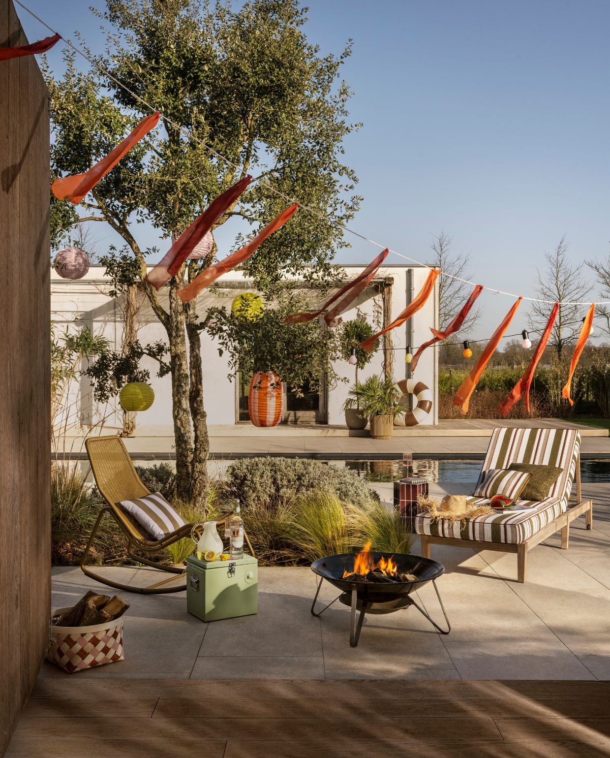 Woood tuinschommelstoel Tom, metaal en kunststof, honinggeel € 159,-, bijzettafel Plu (indoor), keramiek, paars, 30x26x42 cm (bxdxh) € 119,-, Zuiver koelbox Cooler be cute, kunststof en metaal, groen € 79,- (shop.vtwonen.nl). Gommaire ligbed Sunny Copenhagen, teakhout, naturel, 202x70x12 cm (lxbxh) € 1395,-, kussenset ligbed Sunny Copenhagen, outdoor stof en sneldrogend schuim, inclusief kussen€ 1645,- (gommaire.com). Petites Pommes zwemband Celine, pvc, charleston, 120 cm (Ø) € 50,- (helloarchie.com). Ikea Solar hanglamp Solvinden, kunststof, roze, 30 cm (Ø) € 7,99, solar hanglamp Solvinden, kunststof, geel, 30 cm (Ø) € 7,99, Solar hanglamp Solvinden, kunststof, oranje en roze, 43 cm (Ø) € 8,99, mand Ommjänge, gevlochten vezel, oranje en roze, set van 2 € 19,99, ledlichtsnoer Solvinden, kunststof, multicolor€ 19,99 (ikea.nl). Kwantum tuinkussen Borgia, polyacryl, groen en grijs € 7,50 (kwantum.nl). Zara Home waterkan, borosilicaatglas, geel, € 17,99 (zarahome.com). Karwei wijnglas bloemvorm, roze € 4,99 (karwei.nl). Overig privébezit. vtwonen Zomerspecial 2026 | styling Sanne Ketelaar | fotografie Stan Koolen
