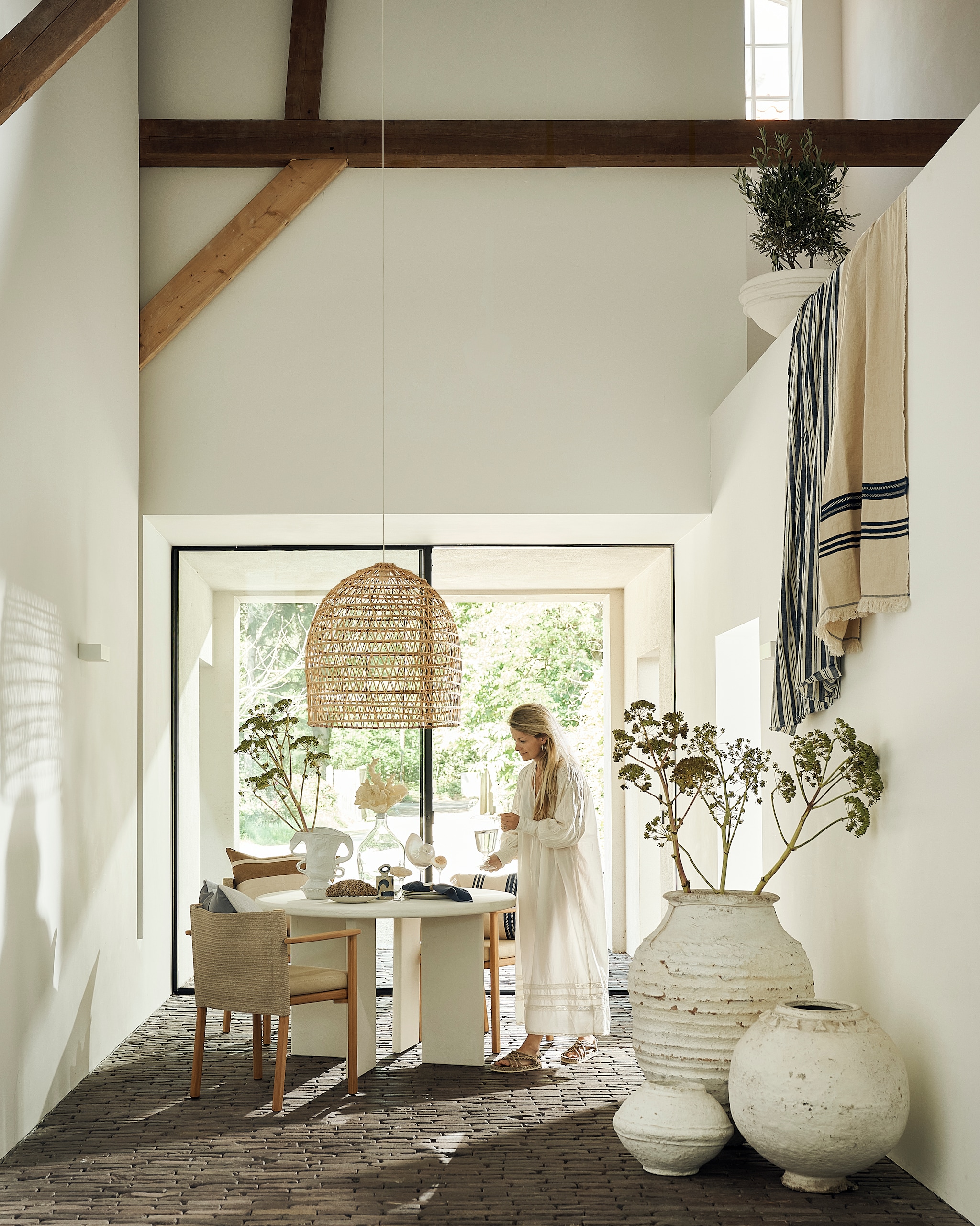 Pebb eettafel, betonlook, shade of linen, 120 cm (Ø) € 1350,- (pebb.nl). Kettal stoelen met kussen Giro, hout en stof, beige, per stuk € 1871,- (dacks.nl). Couleur Locale hanglamp Oum, dadelpalm, naturel € 187,50, Linge Particulier servet, linnen, atlantic blue € 15,-, oude oliekruik Turkije 48, terracotta, wit € 1050,-, vintage graanpot Rajasthan 23, papier-maché, wit € 129,-, vintage graanpot Rajasthan 52, papier-maché, wit € 460,-, kelim kussen, wol en katoen, blauw € 55,-, Serax schaal, papier-maché, wit € 244,-, Jars Céramistes bord Dashi L, keramiek, céladon € 49,- (couleurlocale.eu). Serax opbergpot Sophia 03, keramiek, beige-blauw € 32,-, dinerbord Sophia, keramiek, beige-blauw, set van 2 € 36,-, vaas Rosie 03, keramiek, wit € 120,- (serax.com). Libeco kussenhoes Julian, linnen, railroad stripe € 113,-, kussenhoes The Indigo Stripe, linnen, indigo € 97,-, deken The Indigo Stripe, linnen, indigo € 247,- (libecohomestores.eu). Zara Home gestreepte deken, katoen, wit-blauw € 49,99, kan, borosilicaatglas € 17,99, jurk, katoen, wit € 69,99 (zarahome.com). Maisonnl vaas met nepkoraal, glas, wit € 189,-, schoenen, gevlochten touw, bruin € 89,- (maisonnl.com). Planten en takken, prijs op aanvraag (waterdrinker.nl). Overig privébezit.