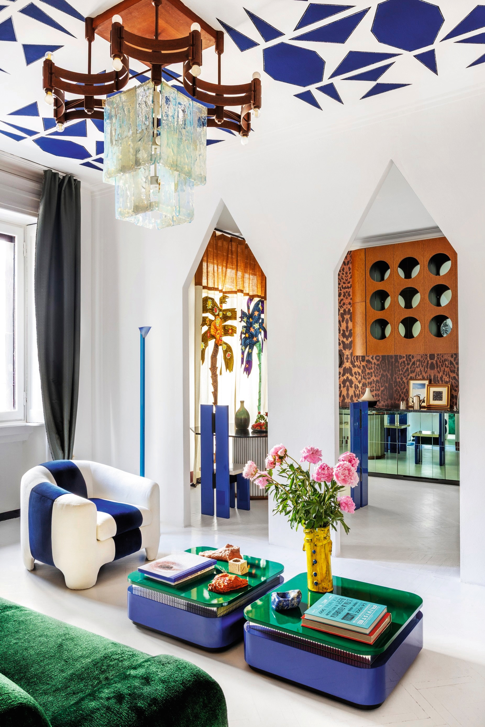 Bank en fauteuil Softhouse Design. Salontafels van Maria Vittoria Paggini voor Softhouse. Gele vaas Giuseppe Palermo. Objecten Tatiana Brodatch. Schilderij Alessandro Florio. Vintage kroonluchter Carlo Nason. Gordijnen met palmbomen Pierre Frey.