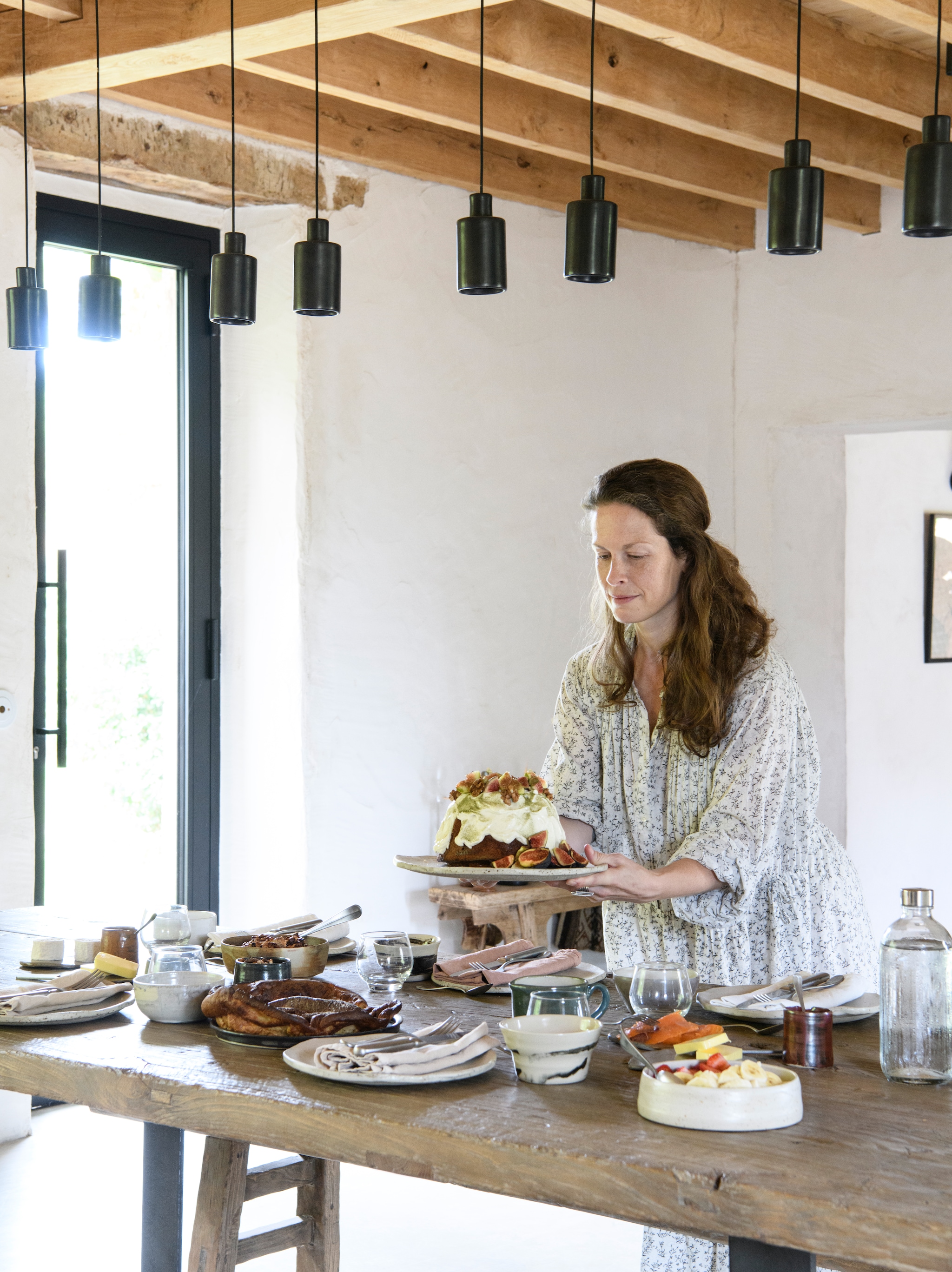Kelly zet haar vijgencake op tafel. Eettafel   Atmosphère D’Ailleurs. Hanglampen David Pompa.  Servies gemaakt door François’ moeder. Glaswerk  van Kelly’s grootmoeder. Servetten Couleur   Chanvre. Wand afgewerkt met natuurlijke   kalkpleister. vtwonen Landelijk 09-2026 | styling Inge van Lieshout | fotografie Louis Lemaire | productie Insidehomepage