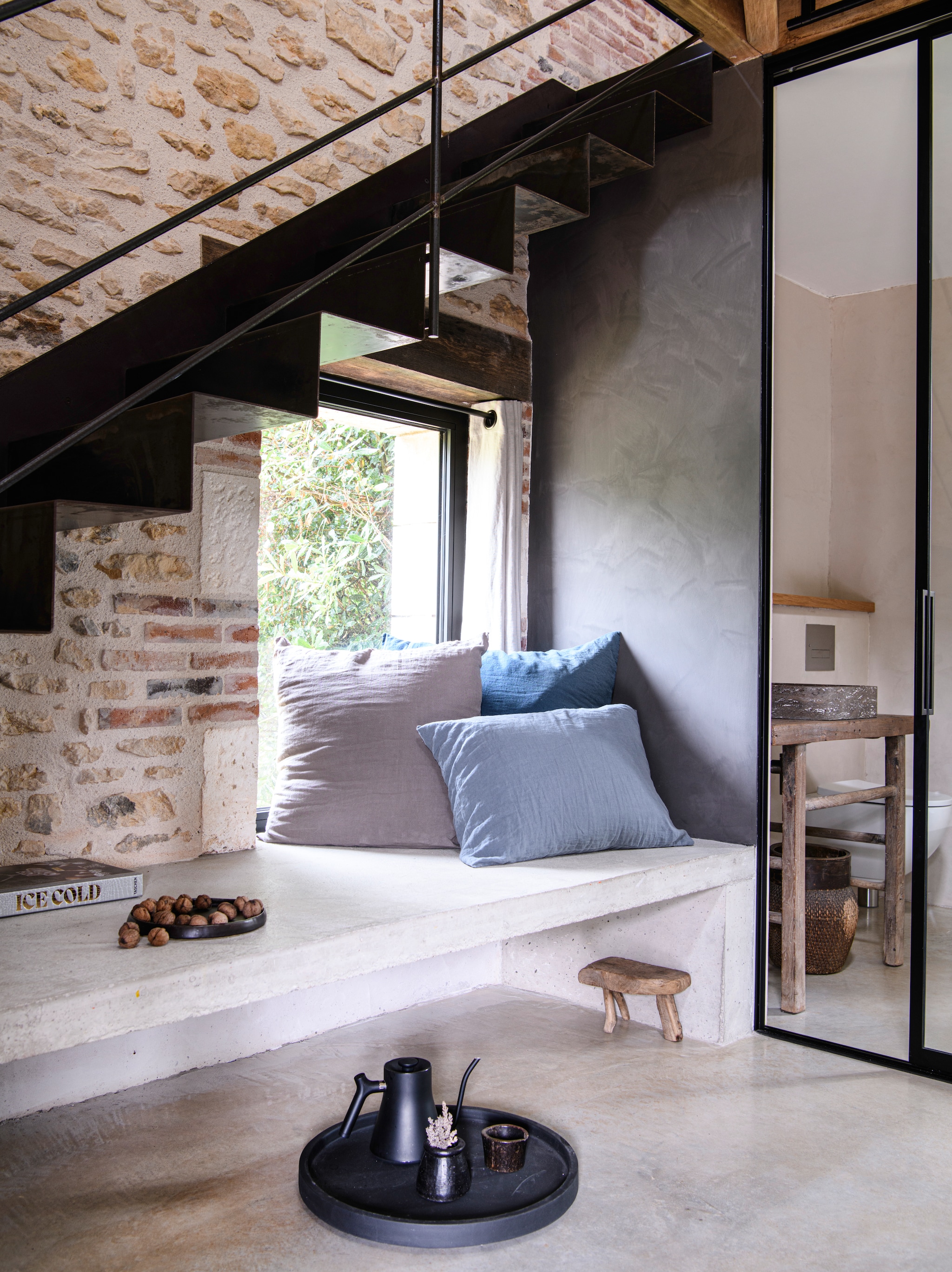 Gastenkamer van de B&B met het bed op de mezzanine. Rechts daaronder de badkamer. 
Op maat gemaakte trap van  onbehandeld staal. Kussens Couleur Chanvre. Op maat  gemaakte glazen deur van  3x4 meter met stalen kozijn naar eigen ontwerp. Dienblad  Bea Mombaers voor Serax.  Waterkoker Fellow. Muur  kalkverf in kleur Réglisse van Ressource. Muur in de badkamer gecoat met natuurlijke kalk- pleister in kleur EF08 van  Ressource. vtwonen Landelijk 09-2026 | styling Inge van Lieshout | fotografie Louis Lemaire | productie Insidehomepage