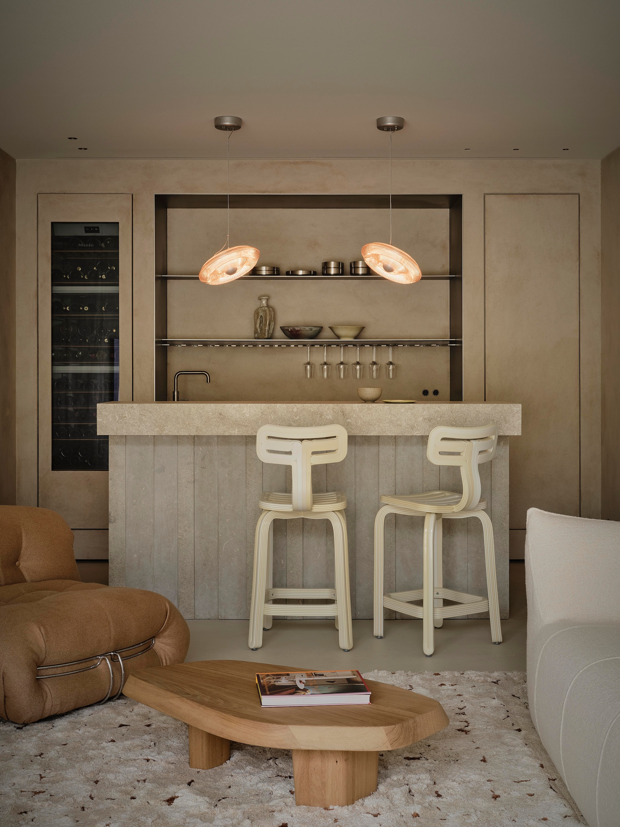 Salontafel maatwerk door Behout, ontworpen door Anne Claus. Vloerkleed Jov. Linker fauteuil Soriana van Cassina. Rechter fauteuil Le Bambole van B&b Italia. Barkrukken en hanglampen Dirk van der Kooij. Keuken en bar maatwerk, ontworpen door Anne Claus. Bar uitgevoerd in Portugese natuursteen. vtwonen design 8-2026 | fotografie Space Content Studio