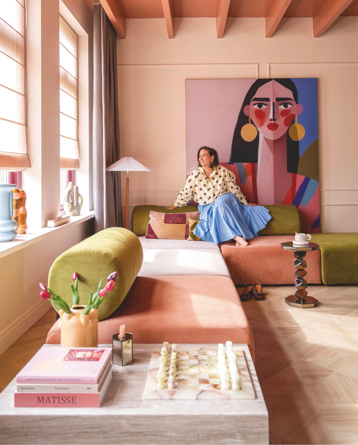 Bank Zitmaxx. Schilderij Carla Van Iersel via Werk aan de Muur. Staande lamp H&m Home. Blinds Kendix. Gordijnen De Ploeg. Vloer Moduleo. Hoektafels, blauwe vaas en spiegeltafel By-Boo. Schaakbord The Vintage Atelier. Kandelaars Kwantum. Kussens Sézane. Verfkleuren plafond Light Cherry Parade en wand Peach Pudding van Histor.