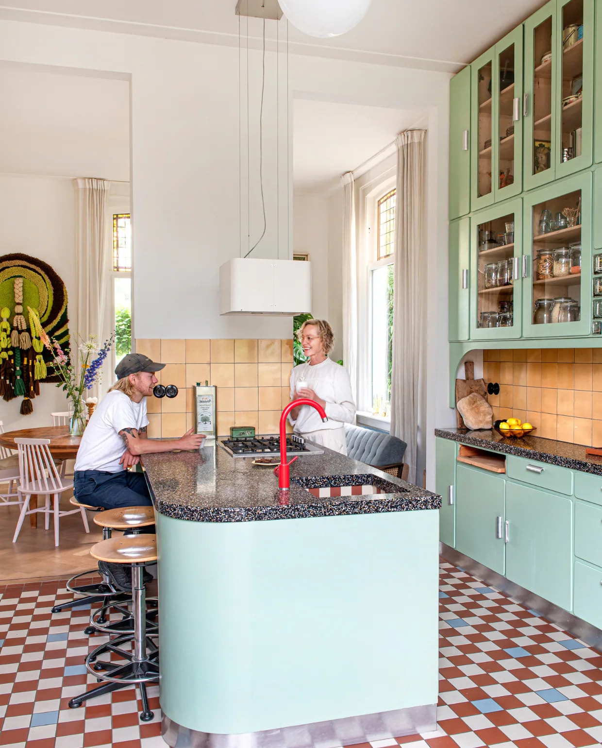 Keuken ontworpen door Studio Daan Hietbrink, verfkleur fronten Room Green 81 van Farrow & Ball. Keukenblad Marcolina Terrazzo en Beton. Rode kraan sanitair.nl. Wandtegels en barkrukken Marktplaats. Vloertegels Winckelmans. Fruitschaal en broodplank uit Frankrijk. Overig onbekend. vtwonen 03-2023 | styling & fotografie Caroline Coehorst i.s.m. Studio Daan Hietbrink
