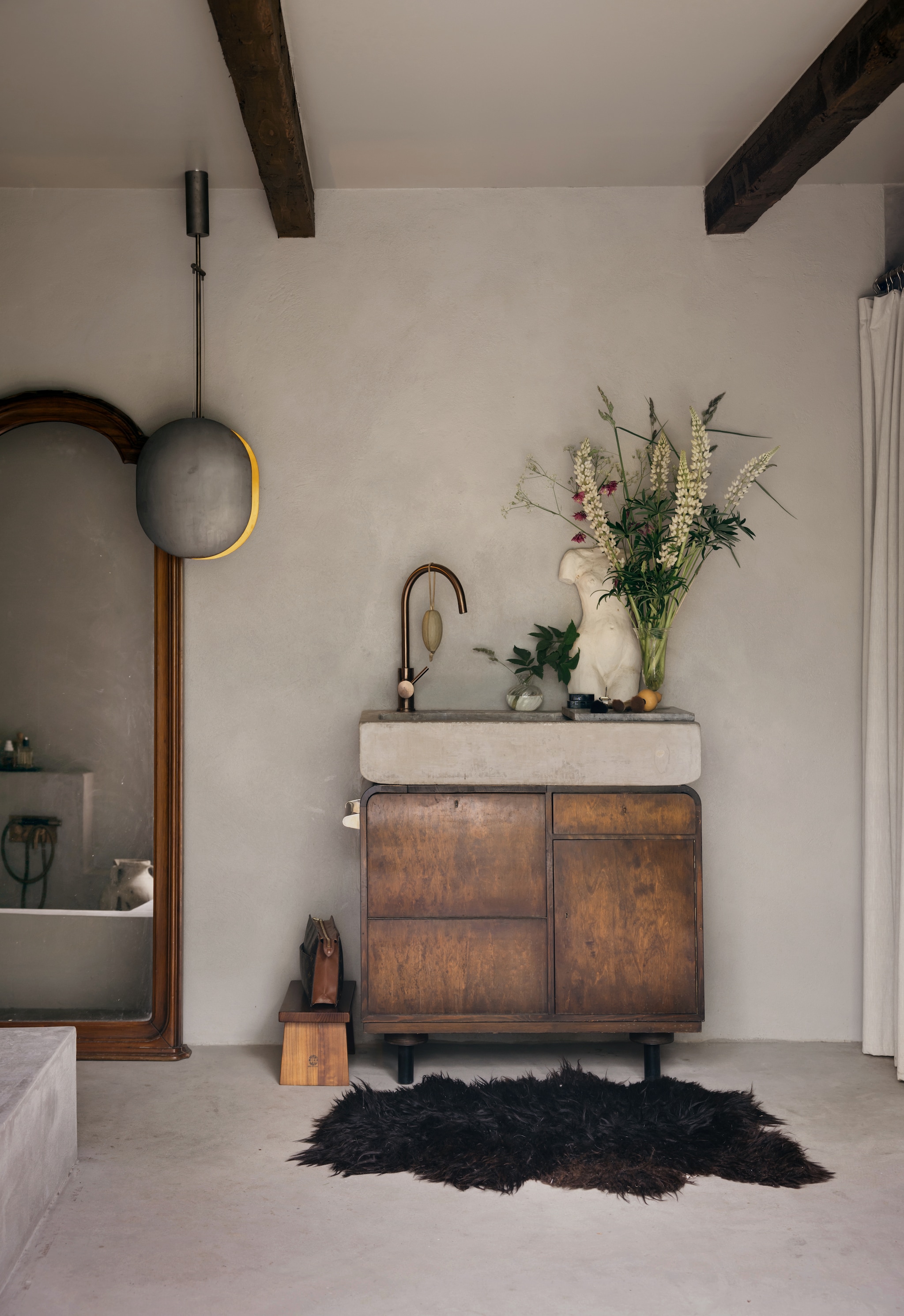 Wastafelmeubel van serveerwagen Nordiska Galleriet uit 1930, met een op maat gemaakte wasbak van beton. Koperen kraan Tapwell. Lamp 101 Copenhagen. Spiegel antiek. Maatwerkbad. Schapenvacht vintage. vtwonen Landelijk 10-2026 | Styling Karolina Vertus | Fotografie Isabell N Wedin | Productie Living Inside