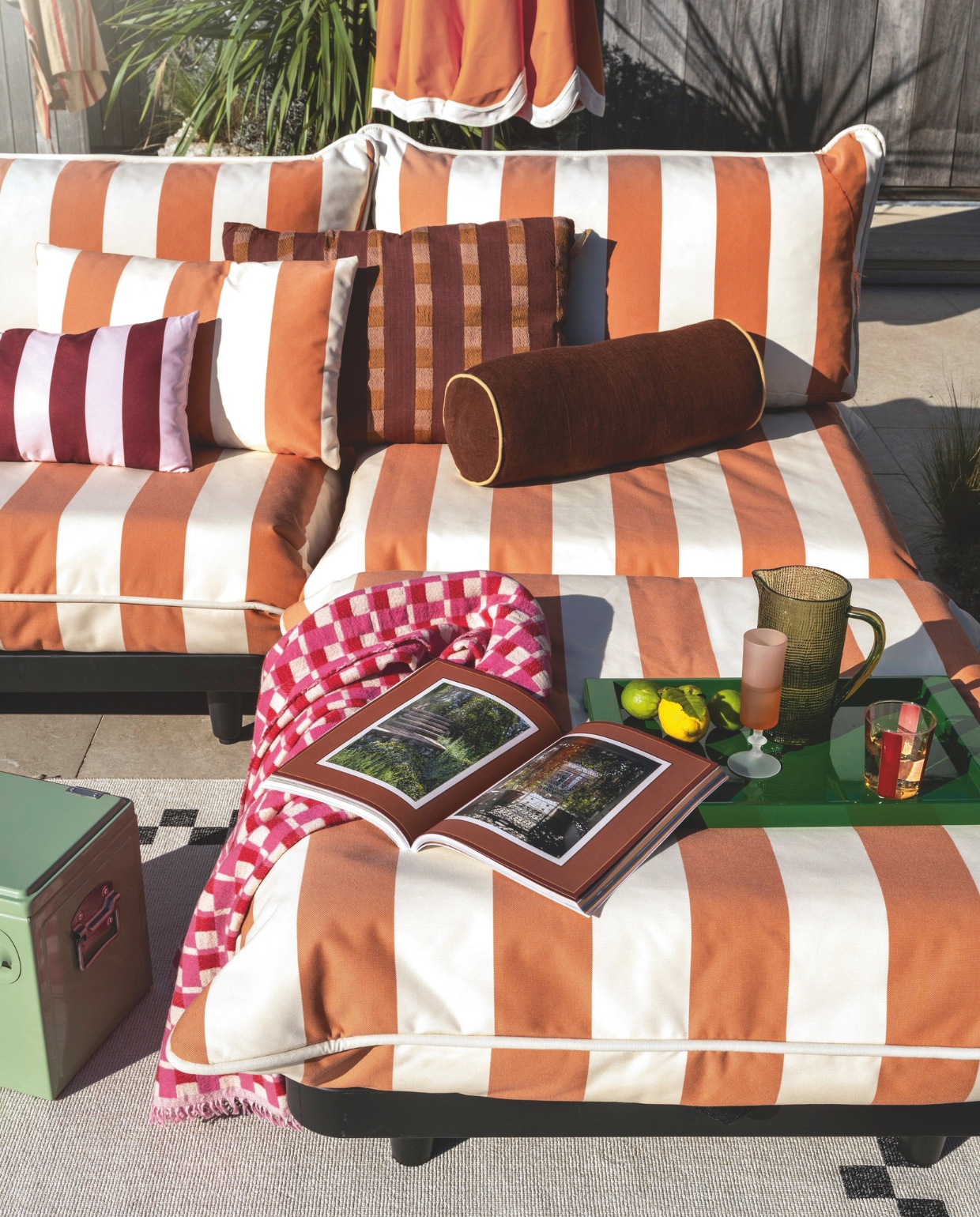 Fatboy loungeset Paletti, aluminium frame en outdoor stof, stripe orange creme, 280x190x90 cm (lxbxh) € 3446,-, Pensol parasol, aluminium en polyester, oranje, 350x350x280 cm (lxbxh) € 499,-, buitenvloerkleed Carpretty grand, polypropyleen, beige en zwart, 300x200 cm (lxb) € 299,-, sierkussen King Pillow, polyester en katoen, beige en oranje gestreept € 55,- (fatboy.com). Zuiver koelbox Cooler be cute, metaal en kunststof, groen € 79,- (shop.vtwonen.nl). Intratuin sierkussen Vienna, polyester, beige en oranje € 19,99 (intratuin.nl). Karwei kussen streep, bruin € 21,99, palletkussen Marleen, textiel, oranje, geel en wit, 120x80x12cm (lxbxh) € 99,99 (karwei.nl). Kwantum loungekussen Lupia, polyester, bordeauxrood/roze € 12,50 (kwantum.nl). Ikea Cilindervormig kussen, textiel, bruin € 16,99 (ikea.nl). Arket dienblad, hout en metaal, groen € 55,-, plaid met print, wol, multicolor, 130x170 cm (lxb) € 99,- (arket.com). Zara Home gestreept glas, rood € 3,99, kan, glas, groen € 25,99 (zarahome.com). Overig privébezit. .vtwonen Zomerspecial 2026 | styling Sanne Ketelaar | Styling assistentie Rosa van Schie, Bente Veld, Eva Otterman | fotografie Stan Koolen