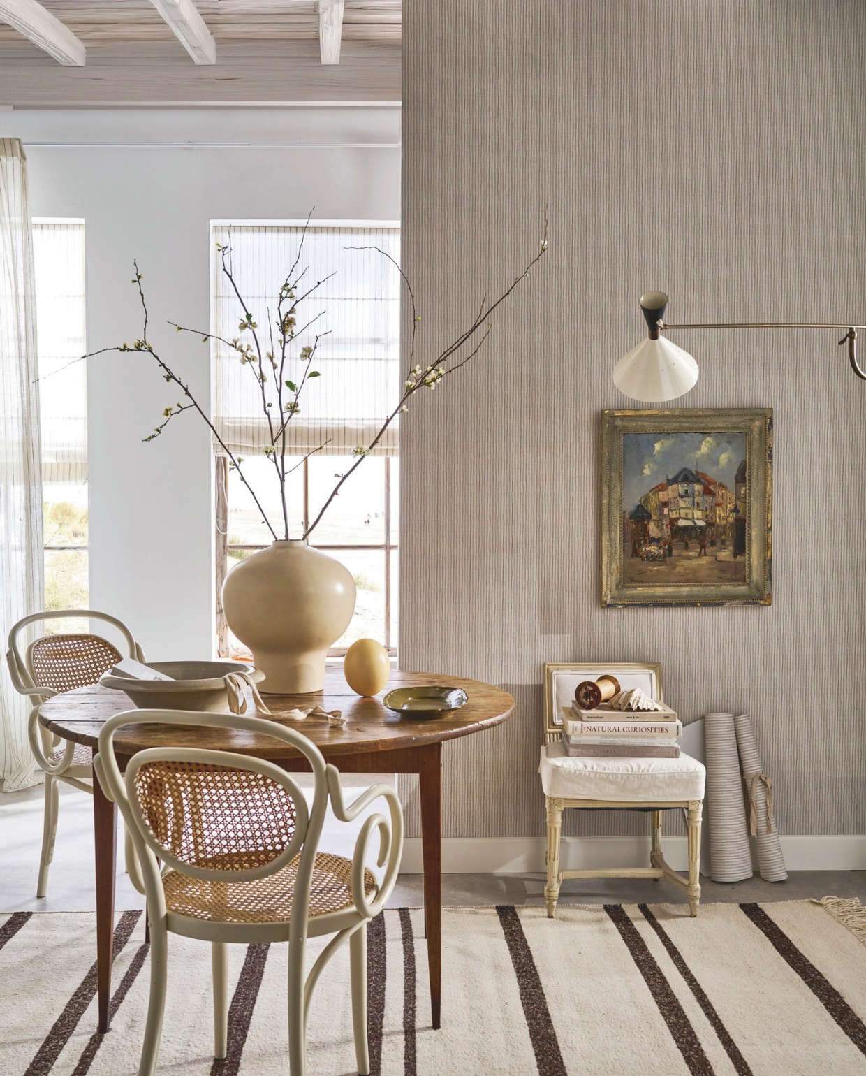 vtwonen behang Double Stripes, vliesbehangpapier, warm grey, 10 m x 53 cm (lxb), per rol € 35,99.M.m.v. prinsheerlijkantiek.nl (schilderij, tafel, gestoffeerde stoel), hkliving.com (vloerkleed), urbannatureculture.com (vaas), karwei.nl (vouwgordijn, muurverf en gordijn van vtwonen), pietjonker.nl (wandlamp). zarahome.com (schaal). Overig privébezit. vtwonen Landelijk 06-2026 | styling Cleo Scheulderman | fotografie Jeroen van der Spek
