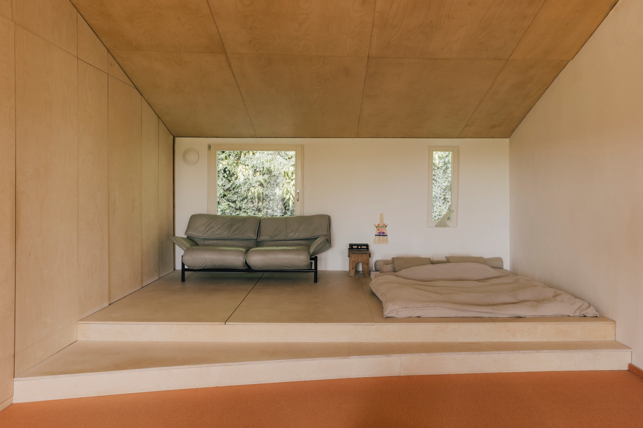 Maatwerkkast, platform en plafond van berkenmultiplex, door Lukas Cober. Bank Veranda van Vico Magistretti voor Cassina. Houten krukje Ikea uit ouderlijk huis. Balinees ceremonieel wandkunstwerk souvenir. Linnen beddengoed. vtwonen design 8-2026 | Fotografie Katoo Peeters