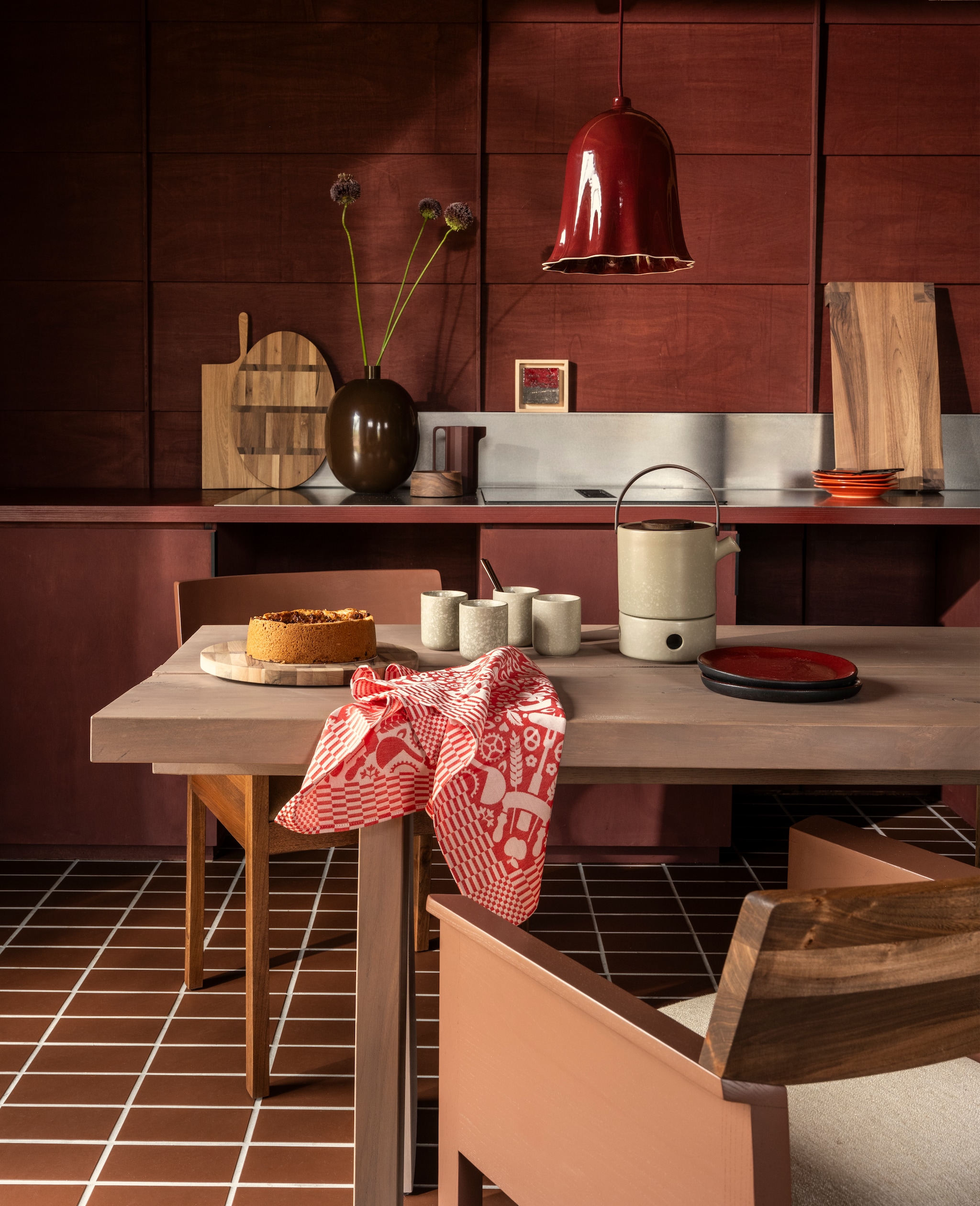 Ateliernvmbr lamp Ciao Bella, keramiek, donkerrood € 269,- (ateliernvmbr.nl). Borden Farmhouse ceramics, keramiek, oranje, set van 2 € 29,95, No2 by Esther kan met oor, 3D-geprint gerecycled hout, bruin € 49,- (no2byesther.nl). Pilat & Pilat stoel Lio, iepenhout, naturel en donkerrood € 1060,-, dienblad Neeltje, eikenhout, naturel € 82,-, snijplank Durk Lutzen ovaal, walnoot, naturel € 75,-, snijplank Fardau, klein, eikenhout, naturel€ 25,-, broodplank Hindrik, walnoot, bruin € 127,-, schaaltje Doppio, walnoot, bruin € 40,-,  (pilat.nl). vtwonen vaas, metaal, chocoladebruin € 69,95, vtwonen bestek Cutlery, metaal, matzwart, 18-delig € 89,-, Bredemeijer theepot met theelicht Umea, keramiek, leemgrijs, set € 84,95, Bredemeijer theemok met onderzetter Umea, keramiek, leemgrijs, set van 2 € 19,95 (shop.vtwonen.nl). Sergio Herman borden, keramiek, rood € 21,50, Cor Unum tegel in lijst, keramiek en hout, roodgrijs/naturel € 55,-(cor-unum.com). Studio Job theedoek Folklore, katoen, rood/wit € 22,-(textielmuseum.nl).  Winckelmans tegels, keramiek, chocolat/brun, 20x10 cm (lxb), per 25 € 60,69 (vd-pol.nl). Wand afgewerkt met Linolie lijnzaadolie, satin woodoil rom, rood, prijs op aanvraag (linolie.dk). Tafel privébezit. vtwonen 10-2026 | styling Marianne Luning | fotografie Stan Koolen