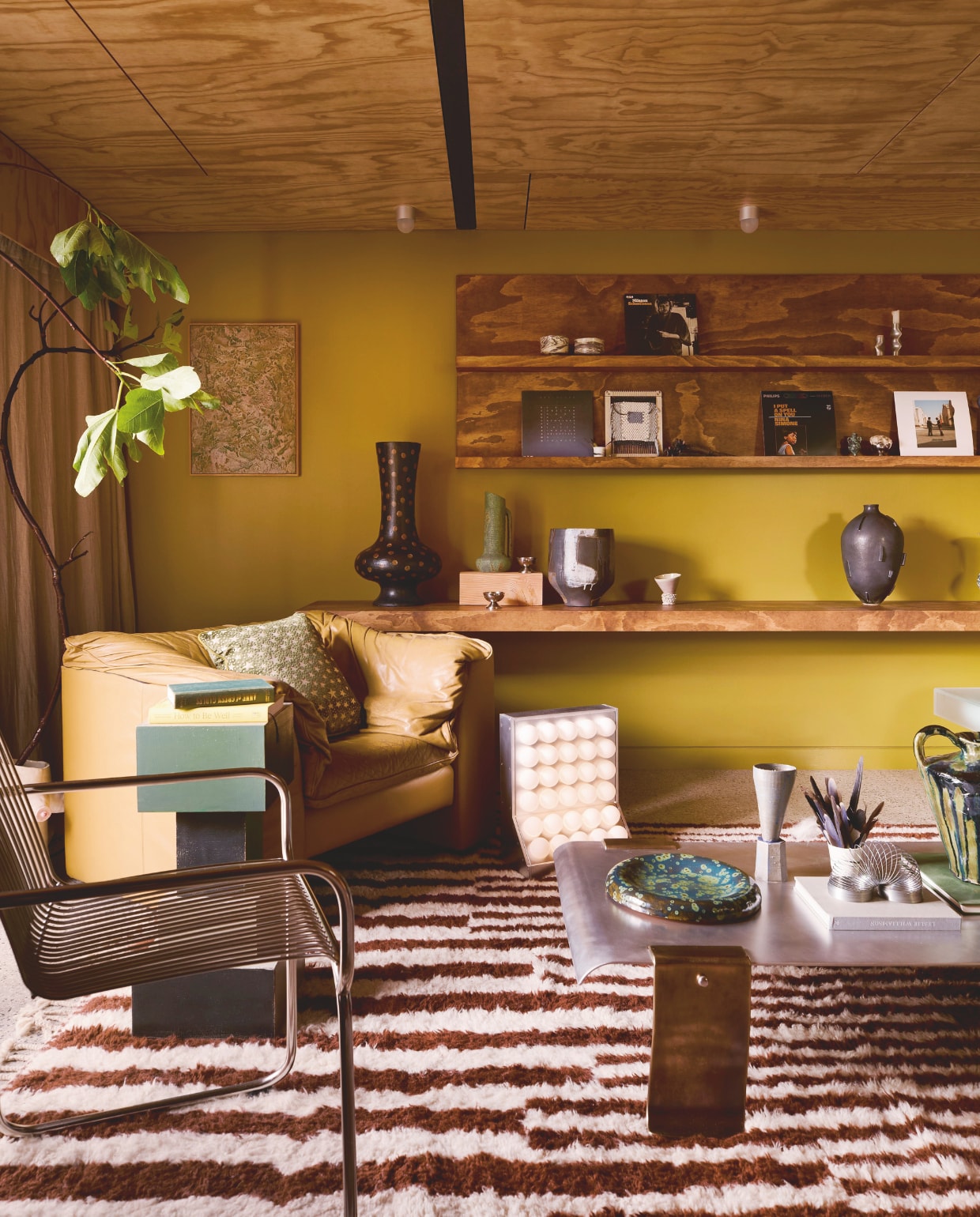 Muurkleur Plantation Island van Dulux. Radiator Ply van Maxiply. Plafondlampen Objectsforthought. Kunstwerk links Ro Noonan. Fauteuil vintage uit de jaren 80. Kussen Celeste van Tali Roth. Salontafel Jordan Fleming. Op salontafel: bord Mossy Platter van Rina Bernabei, object Trophy #2 van Alterfacts en sculptuur Drip Urn van Kelly Brown. Vloerkleed Moroccan Shag van Afrikesh op Etsy. Bovenste wandplank: luciferkommetjes Fdo Studio, kandelaar Locki Humphrey. Onderste plank: donkergrijze vaas Astrid Salomon/Bastard Ceramics, hoge vaas Martyn Thompson, groene vaas Tantri Mustika Ceramics, pot Steph Wallace, object Trophy #4 van Alterfacts. vtwonen Trendspecial 2026 | fotografie Martina Gemmola