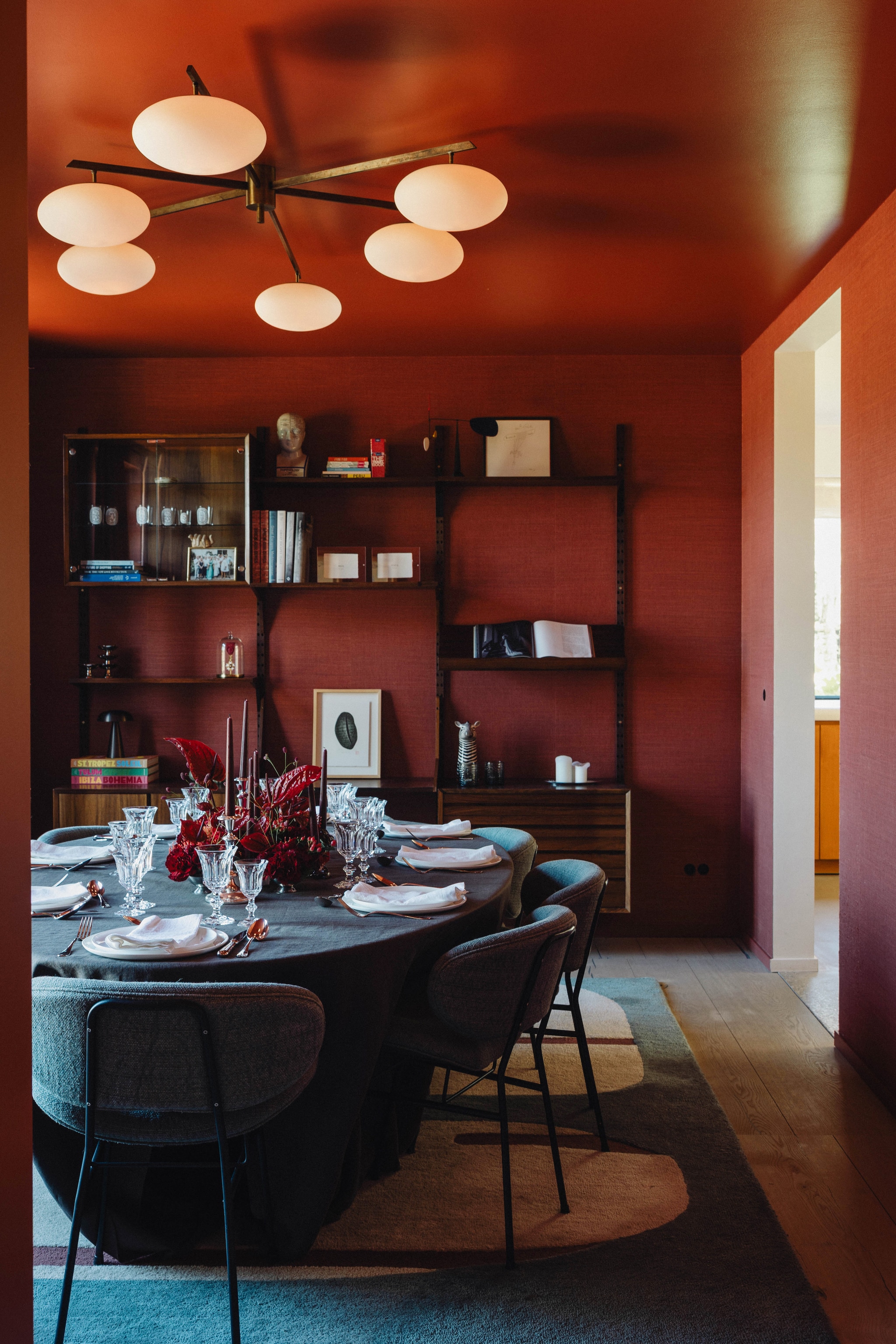 Juten behang Nobilis. Glanzend plafond kleur Picture Gallery Red van Farrow & Ball. Tafellaken en stoelbekleding van stof van Designs of the Time. Tafel Saarinen van Knoll. Vintage stoelen via L’atelier 55 in Saint-Tropez. Vintage plafondlamp Angelo Lelii. Vintage wandmeubel van palissanderhout, ontworpen door Poul Cadovius. Hierop boeken en persoonlijke objecten, zoals de eremedaille van Jo’s grootvader. Vloerkleed Pygmée van Toulemonde Bochart. vtwonen Design 9-2026 | fotografie Stie De Neve